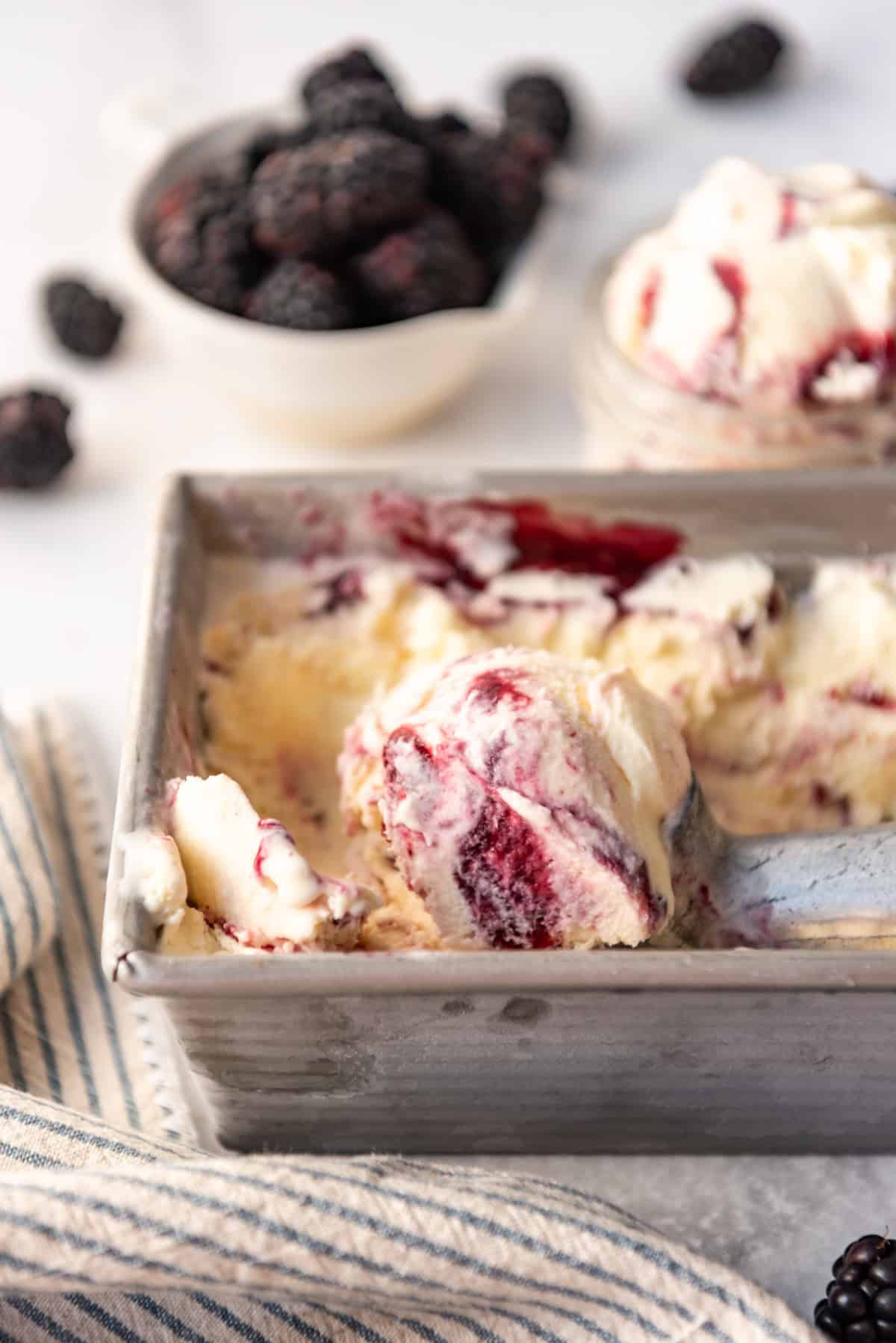 An image of scoops of homemade blackberry swirl ice cream in a container with fresh blackberries behind it.