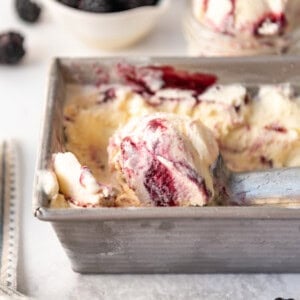 An image of scoops of homemade blackberry swirl ice cream in a container with fresh blackberries behind it.