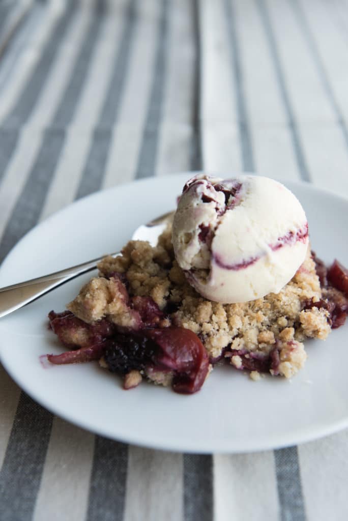 A scoop of blackberry swirl ice cream on top of a fruit crumble on a plate.