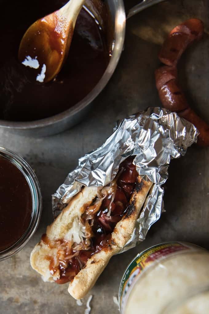 A hot dog topped with J Dawgs sauce and sauerkrat, then wrapped in foil next to a saucepan.