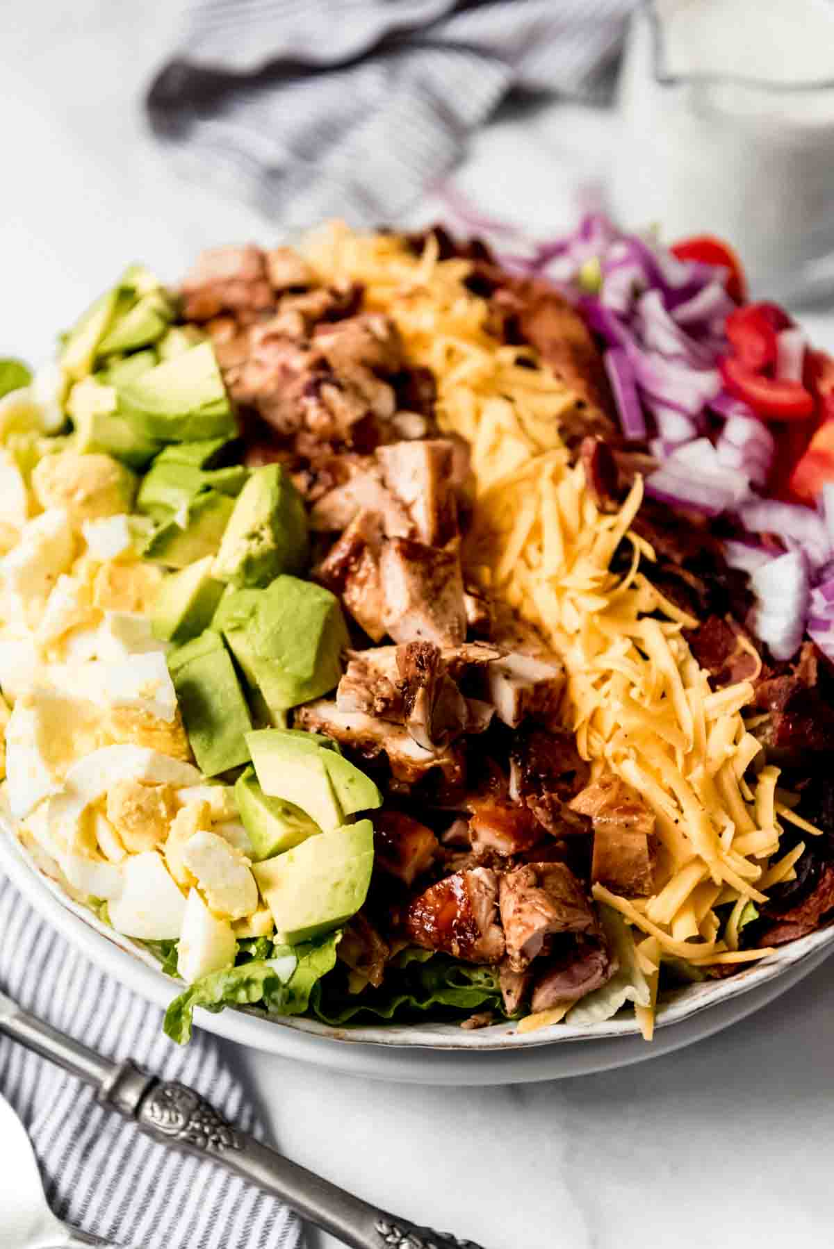 A close image of the top of a bbq chicken cobb salad with hard-boiled eggs, avocado, diced grilled chicken, grated cheddar cheese, bacon, red onion, and tomatoes.