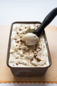A container of homemade toasted almond ice cream with a scoop being scooped out.