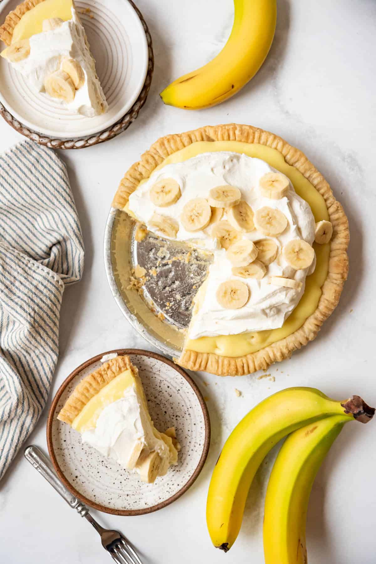 Slices of banana cream pie on plates next to the rest of the pie and some ripe bananas.