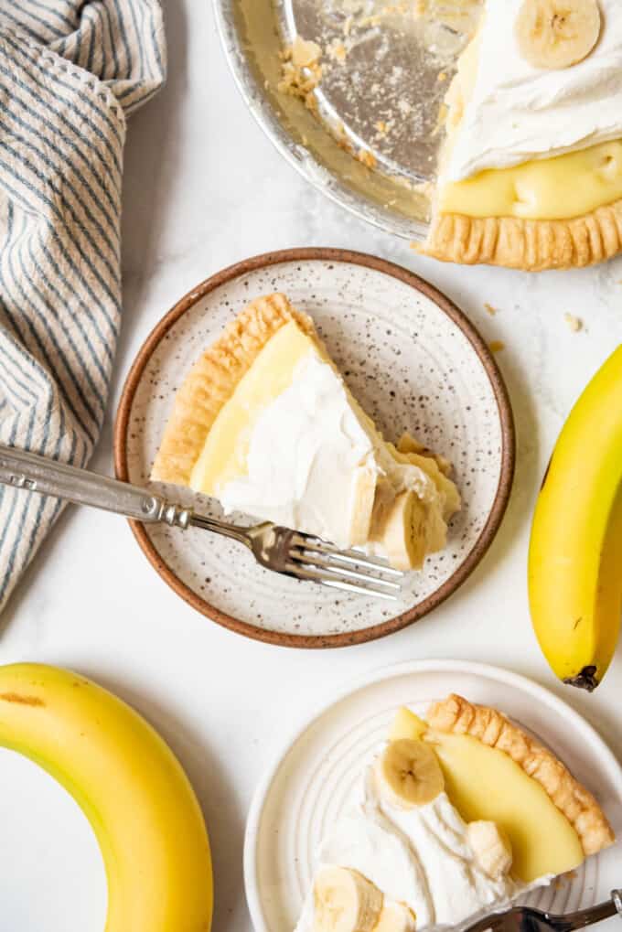 A slice of banana cream pie on a plate next to another slice of pie and bananas.