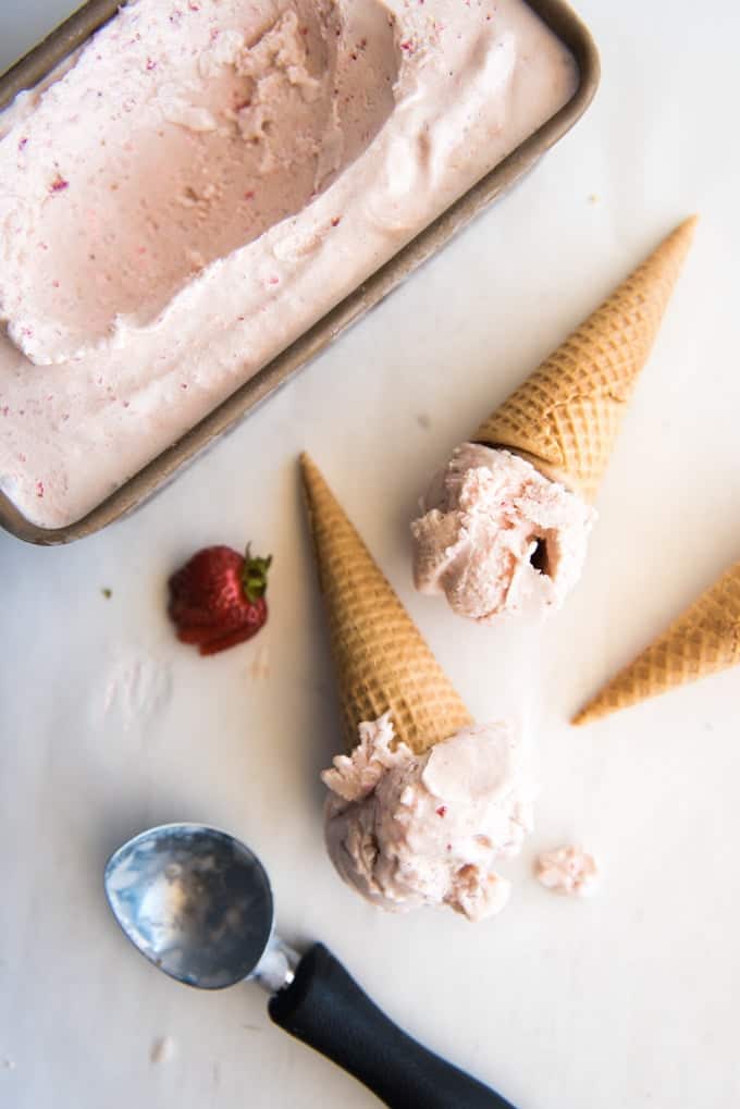 Scoops of homemade strawberry ice cream on sugar cones next to a container of ice cream.
