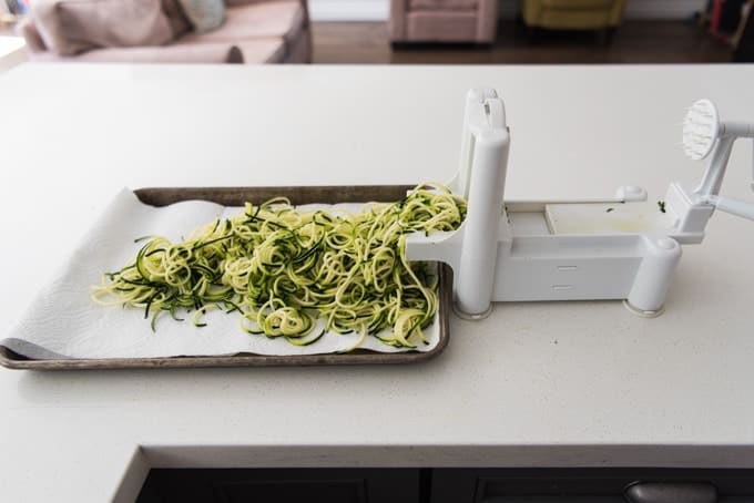 Spiral-cut zucchini "noodles" on a paper towel-lined baking sheet next to a spiralizer.