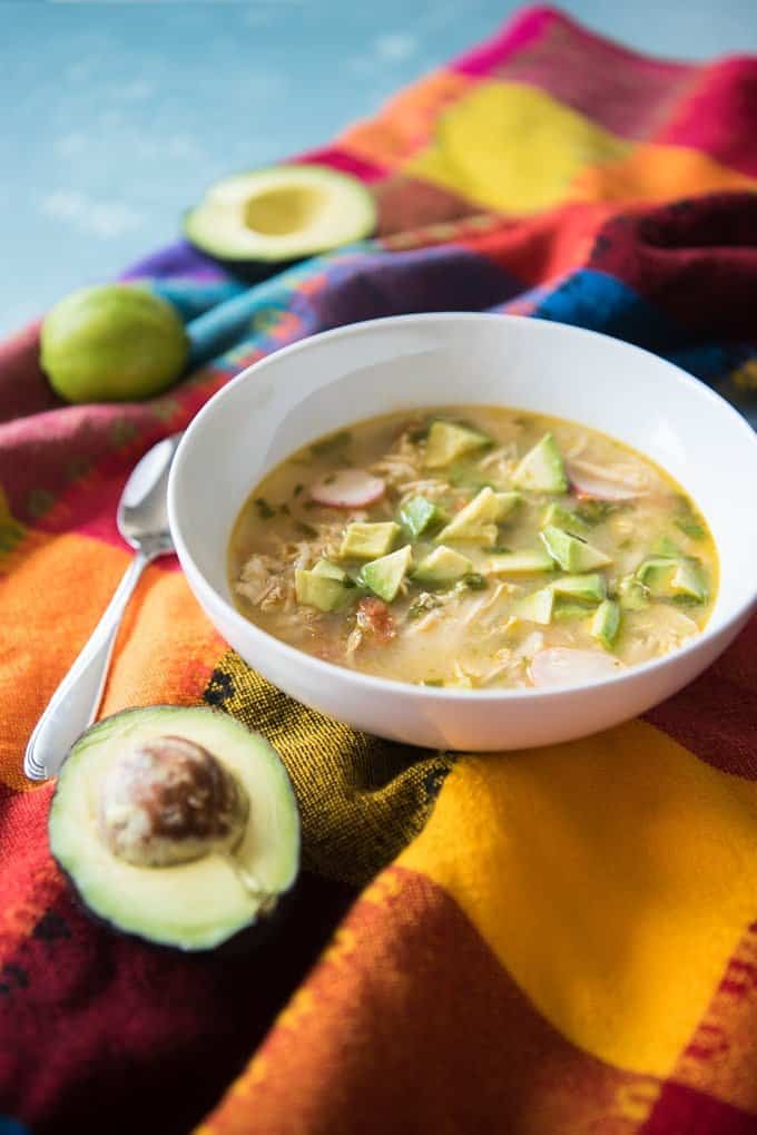 A bowl of chicken soup with avocados and limes on a colorful tablecloth.