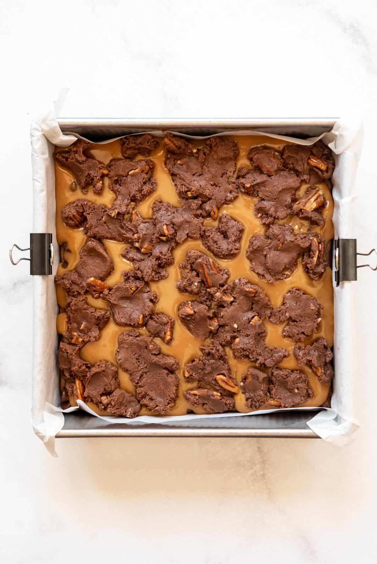 Brownie batter spread on top of a layer of melted caramel in a square baking dish.