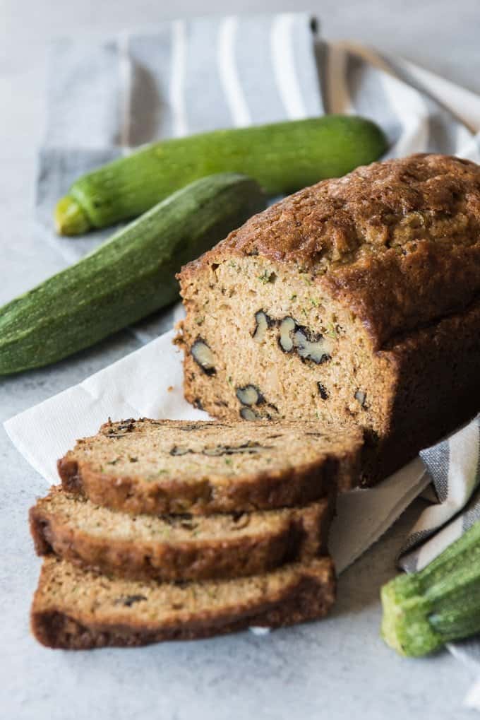 three slices of zucchini bread next to half of a loaf with fresh zucchini in the background