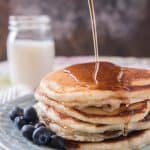 Pouring syrup on a stack ofgolden pancakes with fresh blueberries and a glass of milk to the side