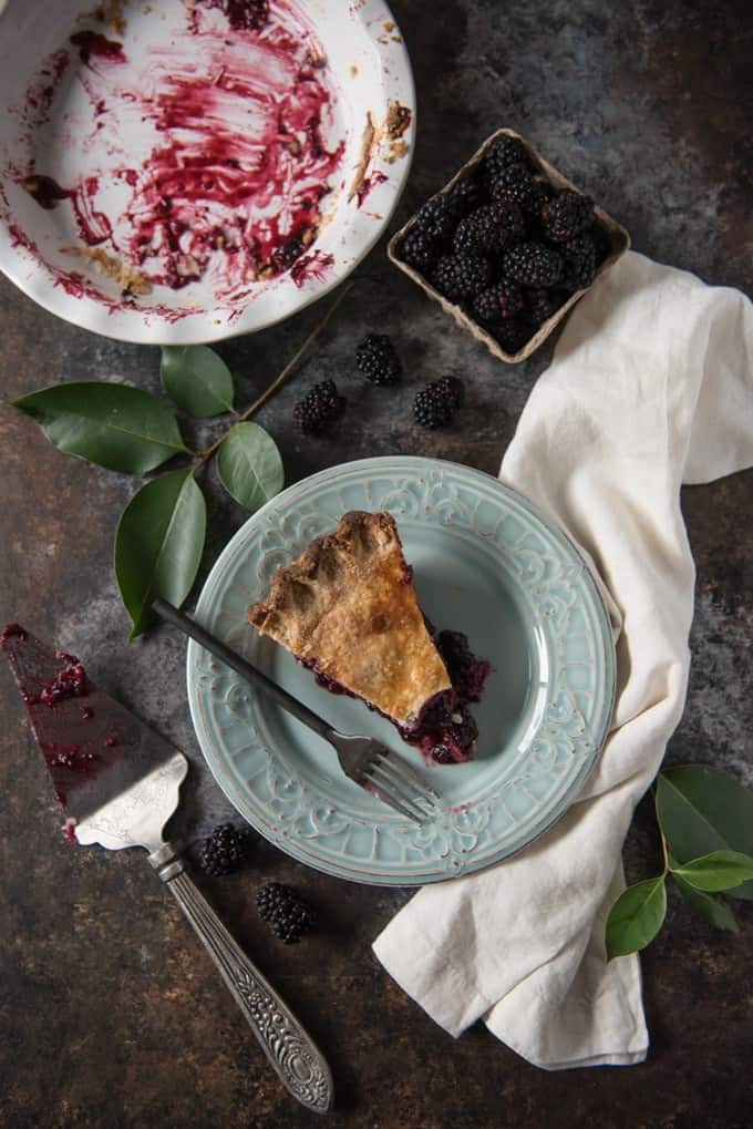 Top view of an empty pie plate with berry juices and a blue plate with a slice of blackberry pie