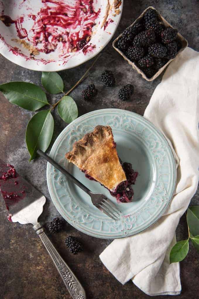 Top view of a slice of pie on a plate with fresh blackberries and an empty pie plate in back.