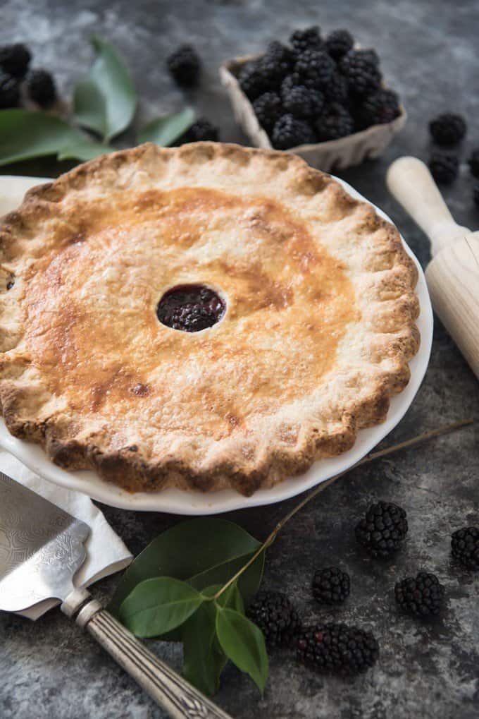 Close up of a baked blackberry pie with fresh blackberries, leaves, a pie server and a wooden rolling pin to the side.