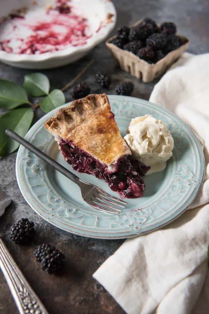 A slice of blackberry pie with a scoop of vanilla ice cream and fresh berries in the back