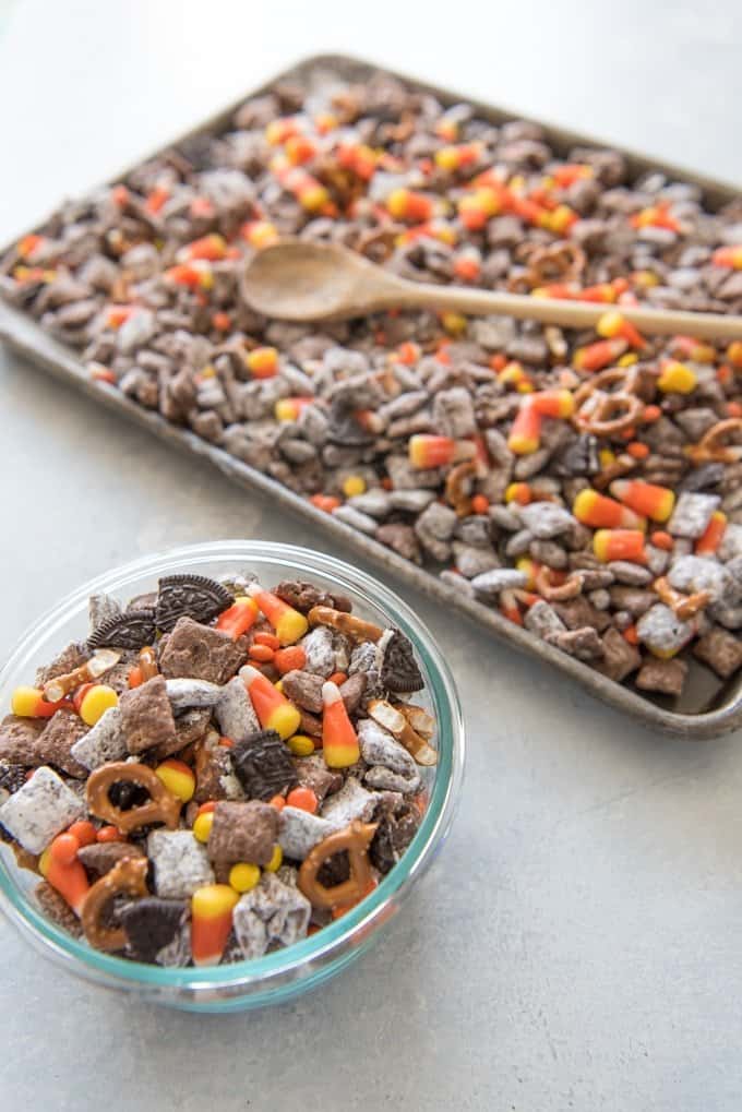 Halloween Chex Mix Muddy Buddies in a bowl andon a baking sheet next to it with a wooden spoon on top