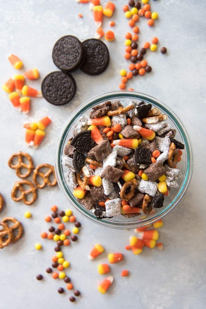 a bowl full of candies and halloween puppy chow mix with scattered candies cookies and pretzels off to the side