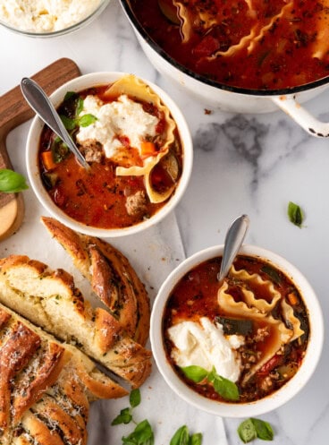 Two bowls of lasagna soup next to a loaf of garlic bread.