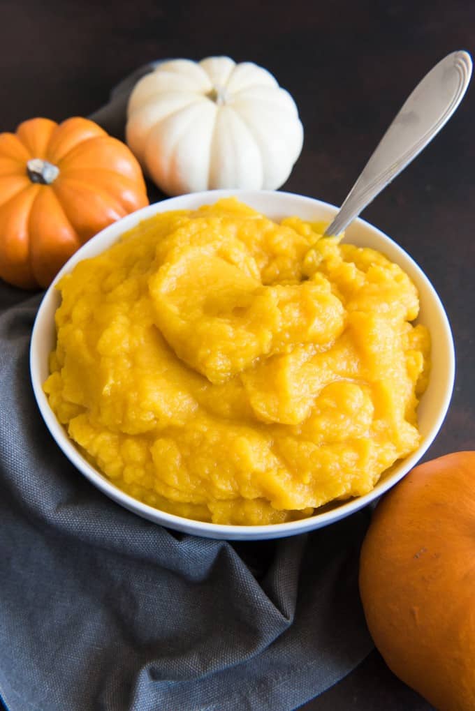 a white bowl of pumpkin puree with a spoon inside and 3 pumpkins and a blue towel around it