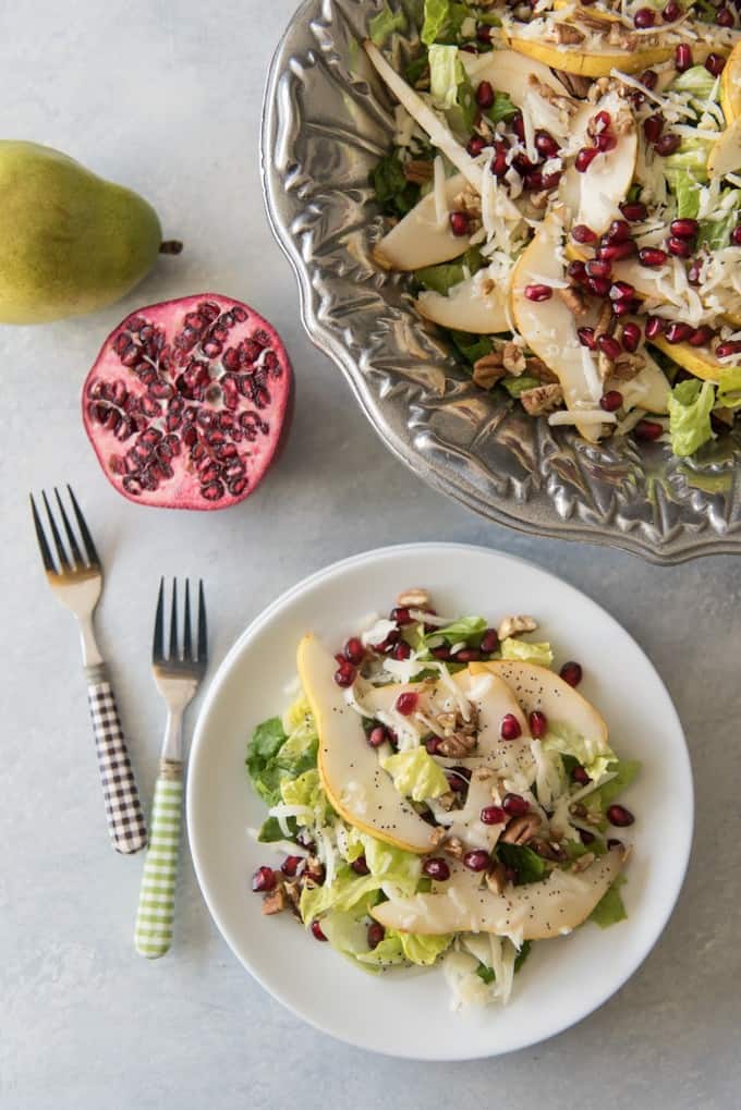 a white plate with a serving of salad and topped with poppyseed dressing with two forks to the side.half a pomegranate and a whole pear to the top next to a bowl of more salad