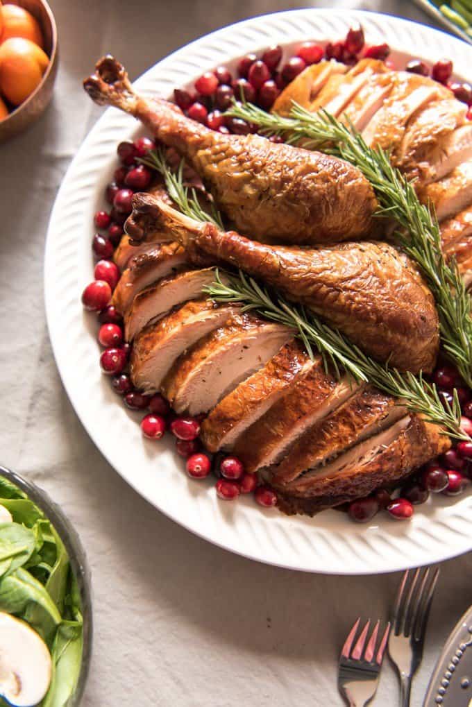 sliced turkey with two legs in the center on a white plate surrounded by cranberries and topped with fresh rosemary