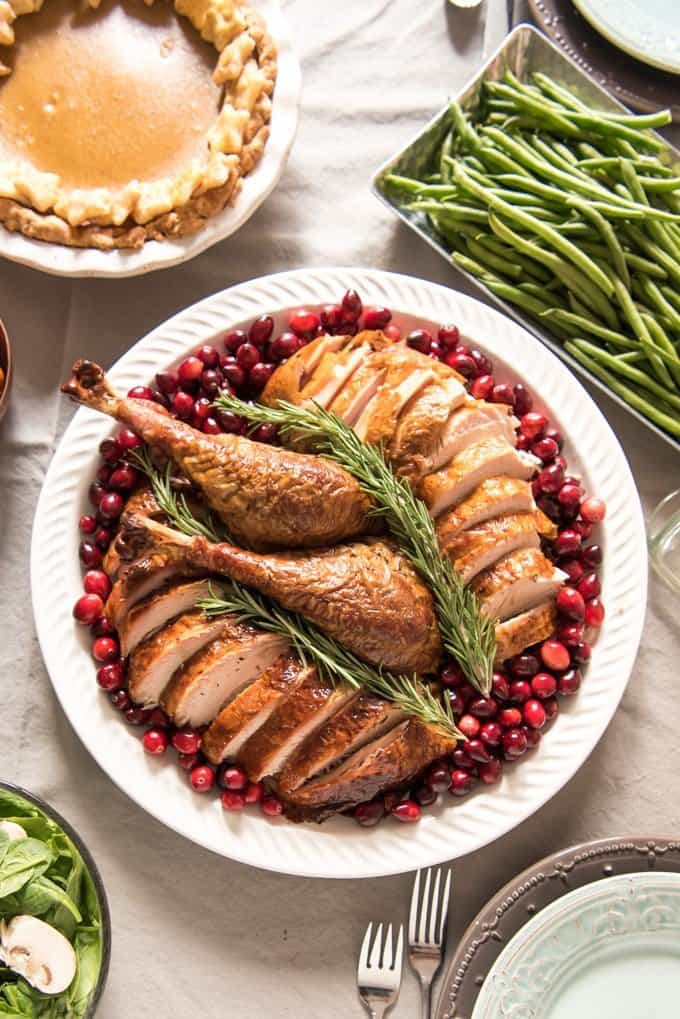 sliced turkey with two legs in the center on a white plate surrounded by cranberries and topped with fresh rosemary a pumpkin pie and some green beans at the upper corners of the image