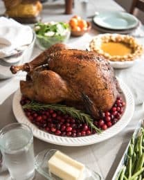 a whole smoked and brined turkey on a white plate with whole cranberries and surrounded by other food dishes