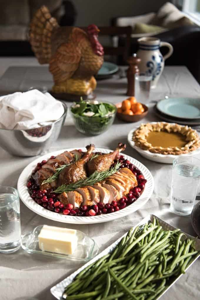 a sliced turkey in a white serving plate with cranberries. Green beans and butter belwo with a pie and other dishes behind as well as a blue and white pitcher and turkey statue
