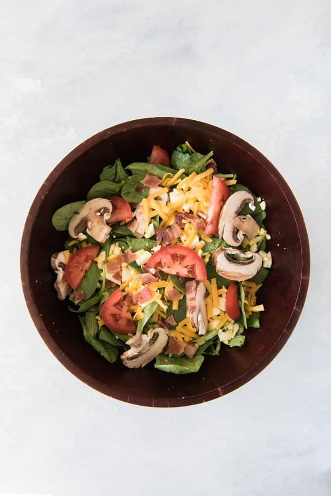 an aerial view of a dark bowl filled with green spinach salad, tomatoes, mushrooms, cheese and more