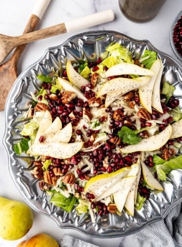 An overhead image of a large salad in a pewter bowl next to serving spoons and two pears.