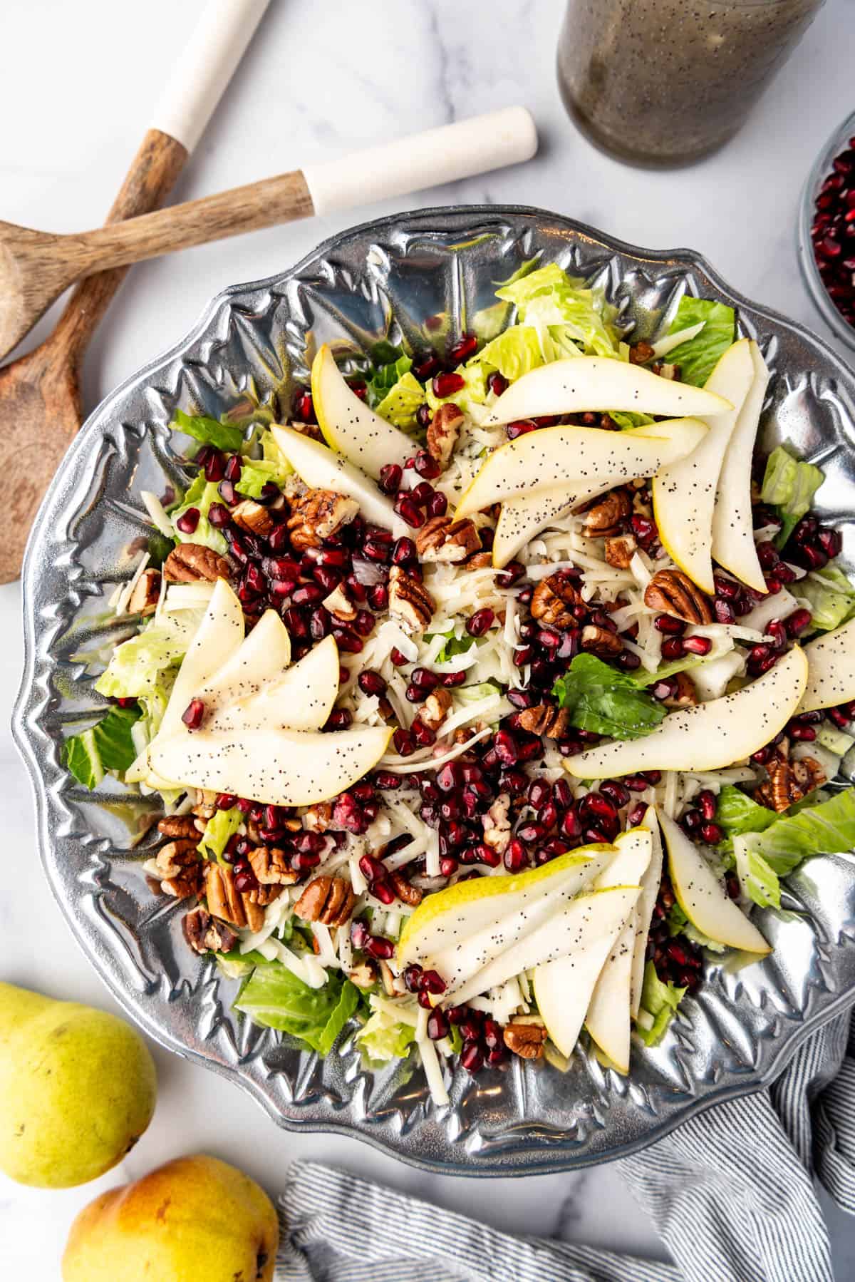 An overhead image of a large salad in a pewter bowl next to serving spoons and two pears.