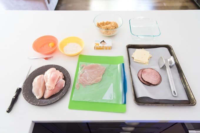 a white counter showing a display of all of the things that you need to make a chicken cordon bleu eggs flour breading chicken breasts meat and cheese one chicken breast is pounded plate already in the center