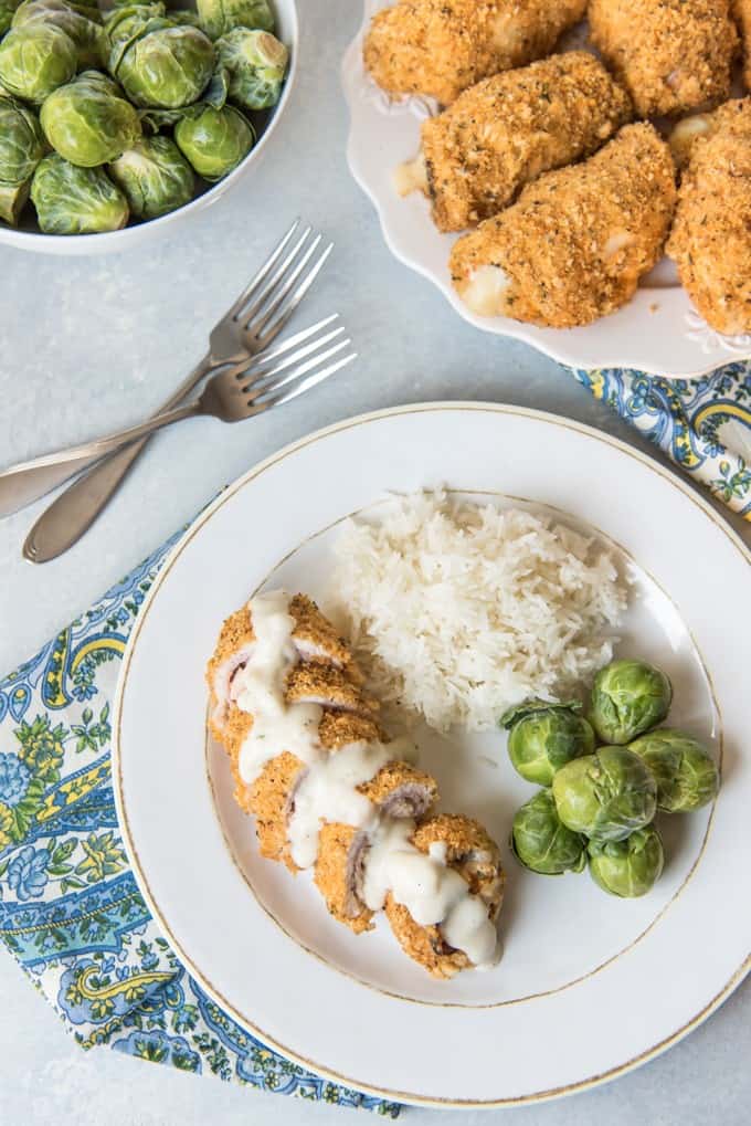 a bowl of brussel sprouts in the top left corner and a plate of baked chicken cordon bleu in the upper right with two forkf resting between them and a white plate with a sliced corndon bleu brussel sprouts and rice