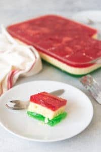 a square of festive jello on a white plate with a spoon next to a baking dish with more jello inside