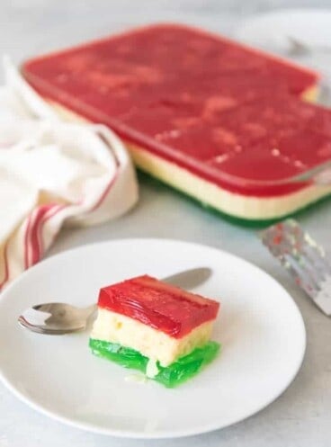 a square of festive jello on a white plate with a spoon next to a baking dish with more jello inside