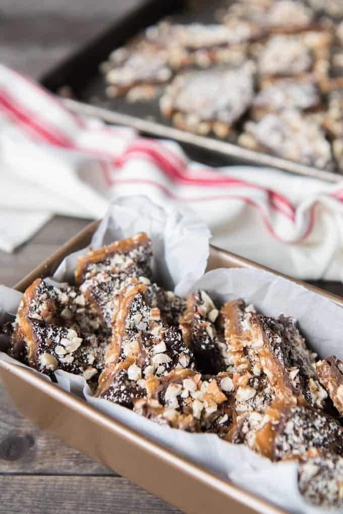 a side angle of english toffee in a bread pan with more pieces on a baking sheet in the background and a red and white striped towel in between