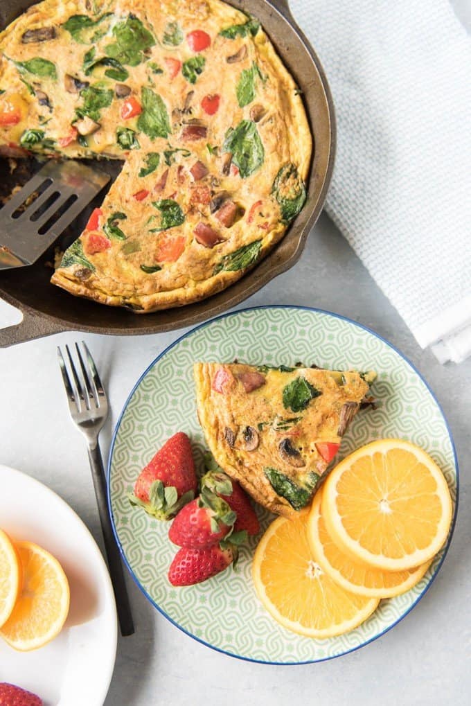 a baked frittata in a cast iron skillet with a serving spatula while a plate in front holds a piece of baked frittat next to sliced oranges and whole strawberries and a fork