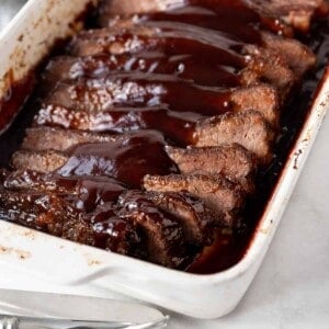 A white baking dish filled with sliced oven roasted barbecue beef brisket topped with a hickory brown sugar bbq sauce.