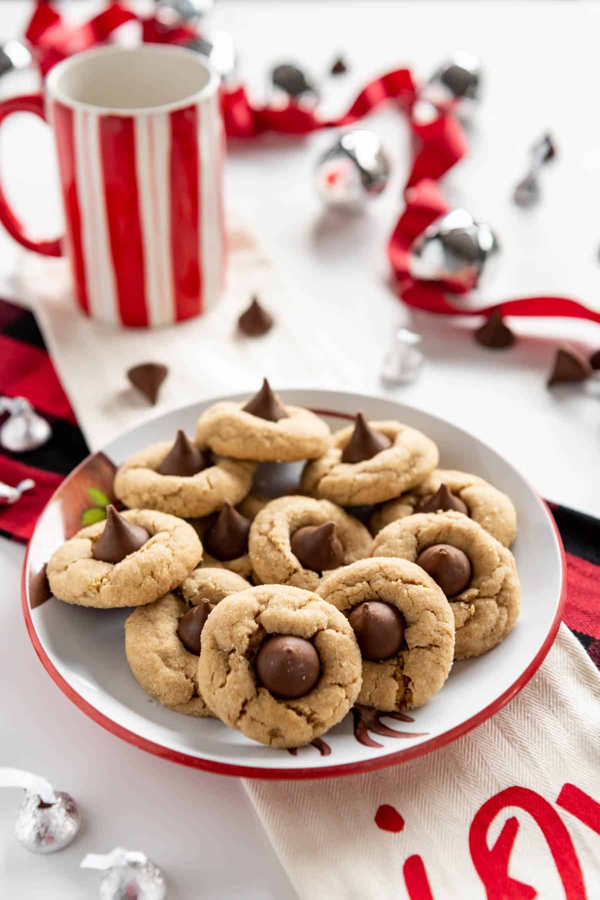 A plate full of peanut butter blossom cookies on a table with red and white decorations and mugs around it and some Hershey kisses both wrapped and unwrapped scattered on the table.