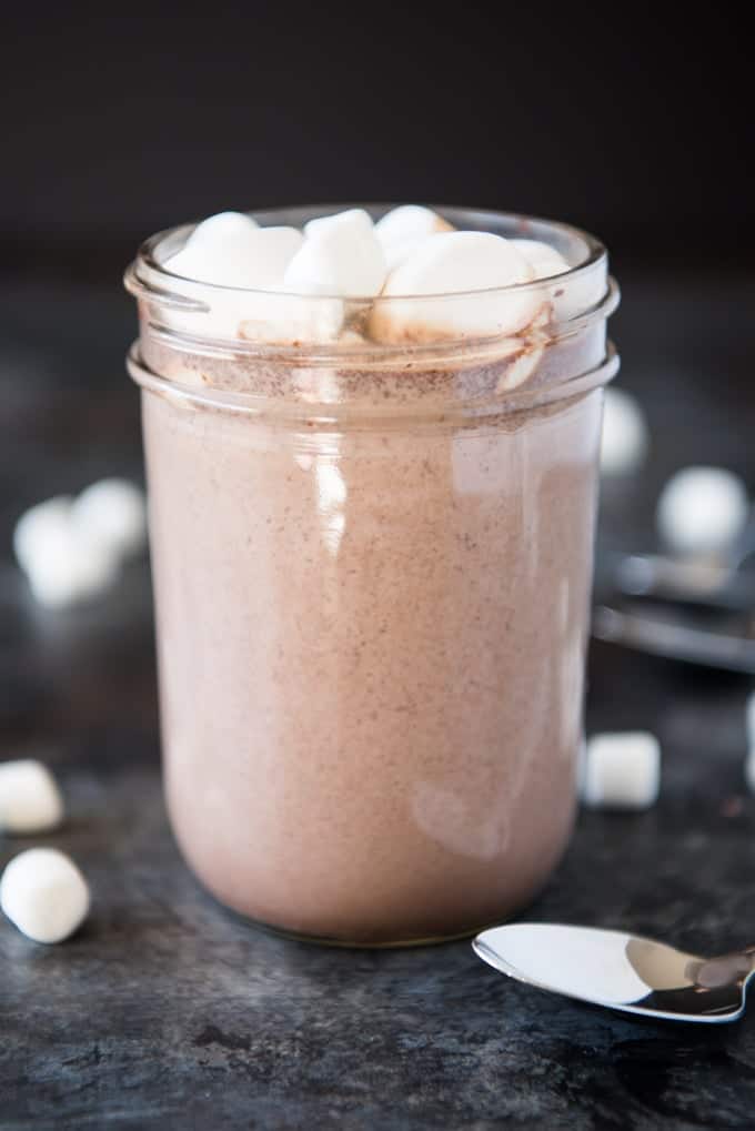 a side view of a jar of cocoa topped with marshmallows with scattered marshmallows around it and a spoon peeking in the front of the image