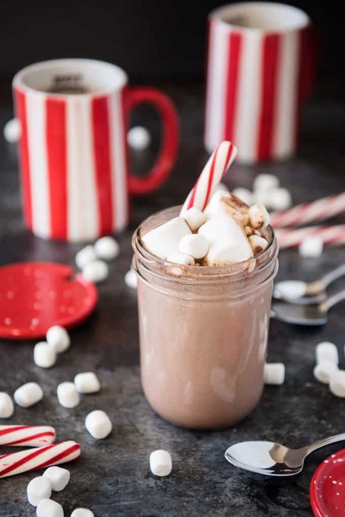 a mason jar of cocoa topped with large and small marshmallows and a candy cane with red and white striped mugs in the backgroun and more scattered spoons marshmallows and candy canes around it