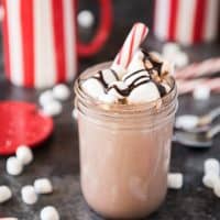 a small mason jar filled with hot chocolate and toppings with mugs and scattered peppermint candies and marshmallows around it