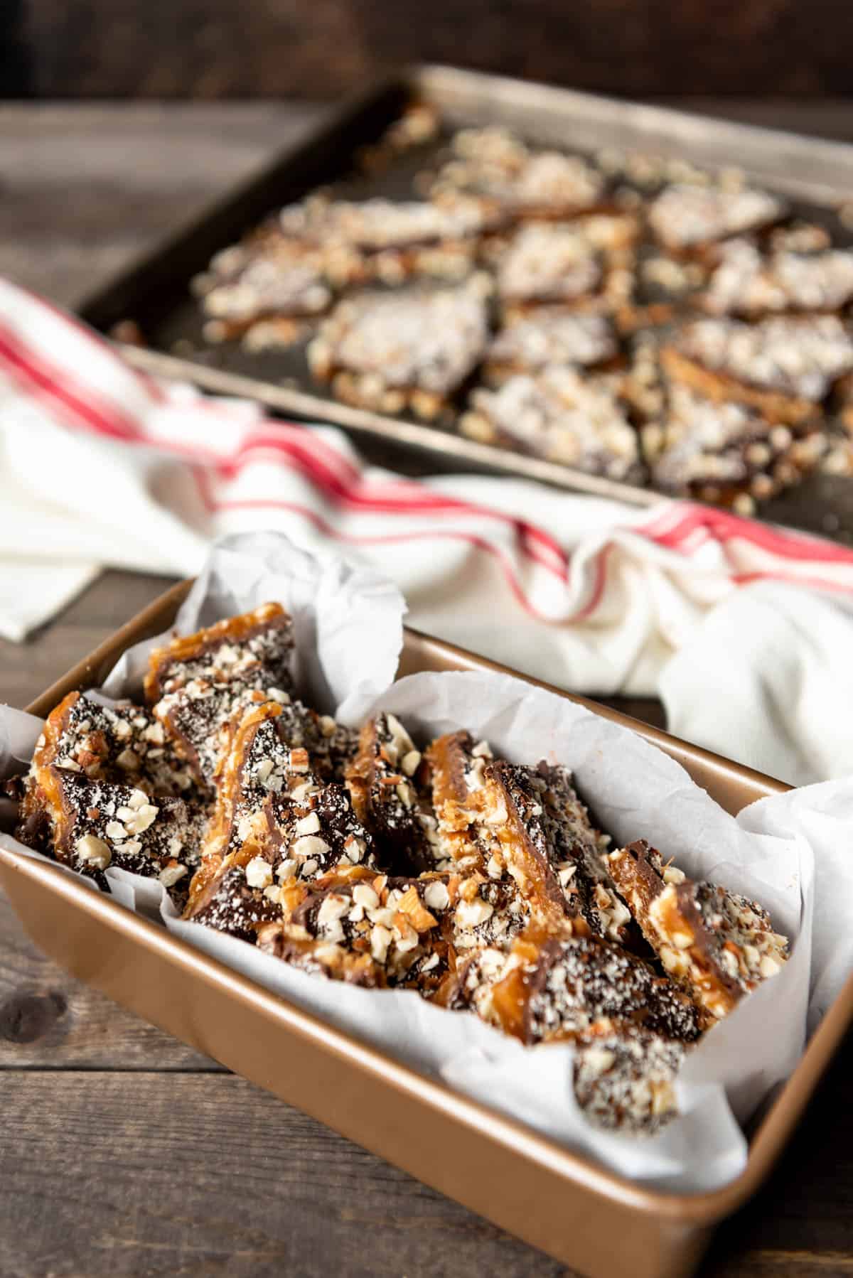 a side angle of english toffee in a bread pan with more pieces on a baking sheet in the background and a red and white striped towel in between