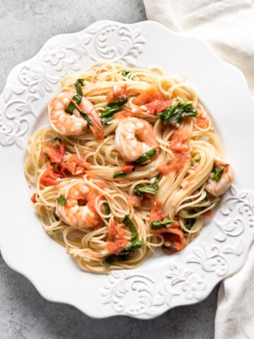 A white plate full of angel hair pasta with shrimp, tomatoes, and fresh basil.