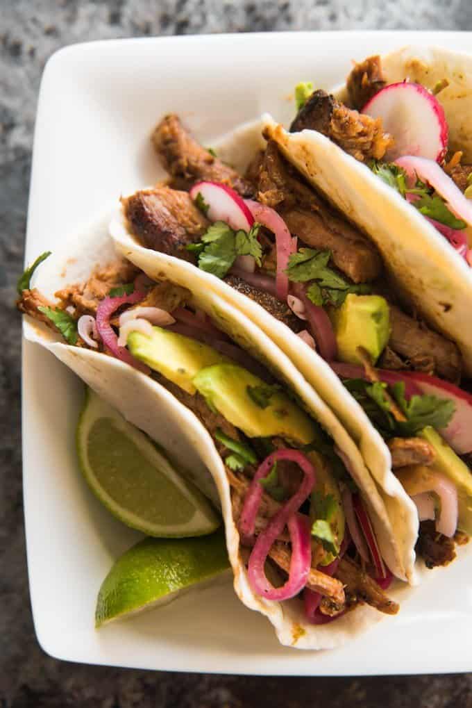 three pork carnitas with garnishes on a white plate next to lime wedges