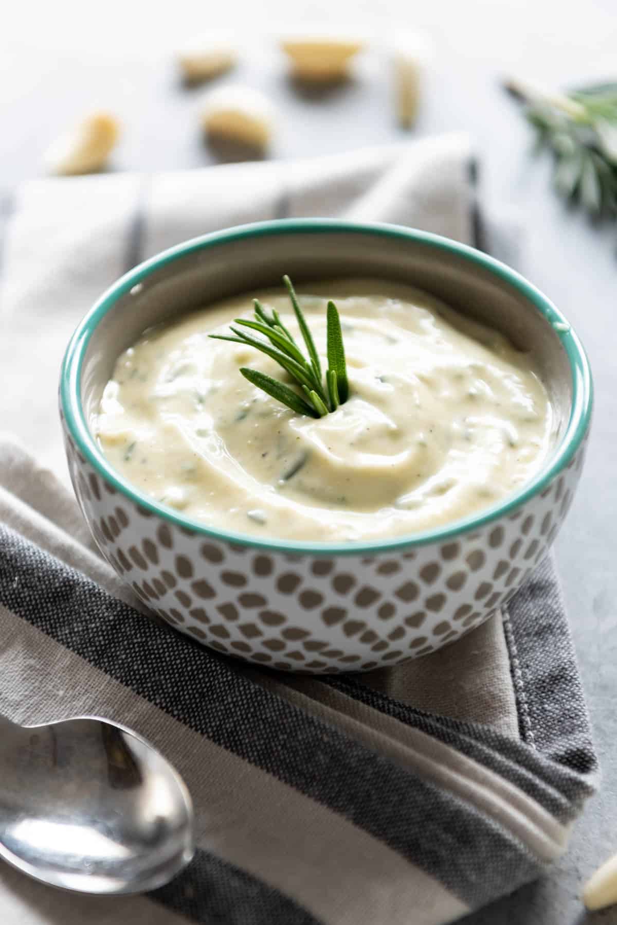 A bowl of garlic and rosemary aioli on a gray and white striped towel with a spoon and garnished with fresh rosemary.