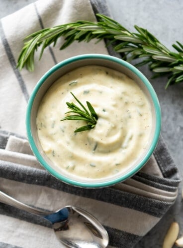 An aerial view of a bowl full of garlic and rosemary aioli on a towel next to a spoon and a sprig of fresh rosemary.