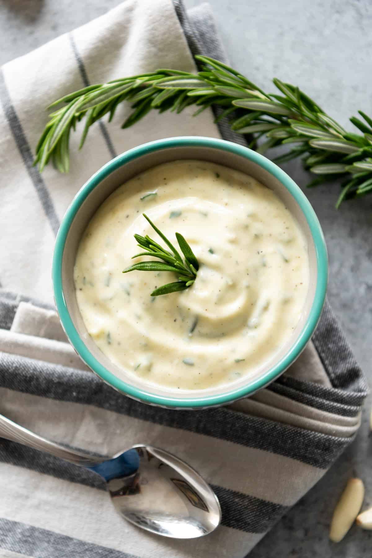 An aerial view of a bowl full of garlic and rosemary aioli on a towel next to a spoon and a sprig of fresh rosemary.