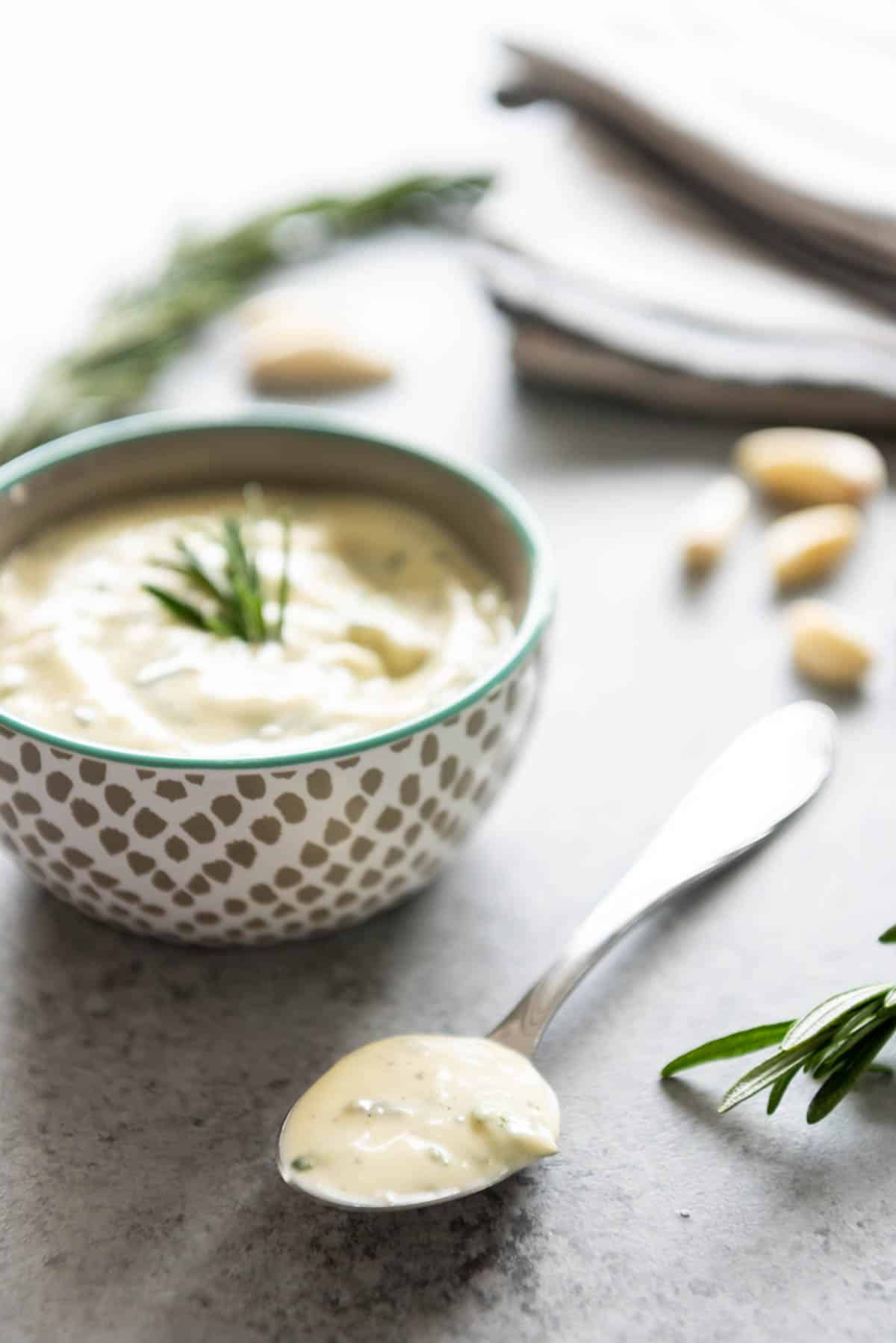 A bowl and a spoon filled with garlic and rosemary aioli with fresh garlic cloves and rosemary scattered around.