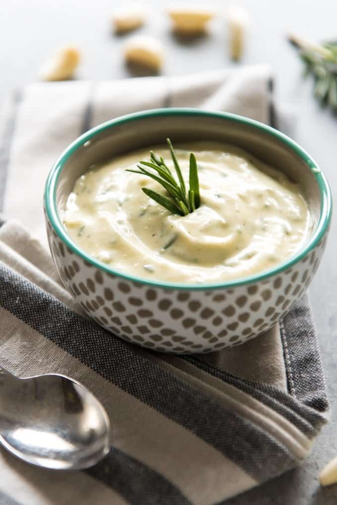 a bowl of garlic and rosemary aioli on a gray and white striped towel with a spoon and garnished with fresh rosemary