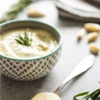 a bowl and a spoon filled with garlic and rosemary aioli with fresh garlic cloves and rosemary scattered around
